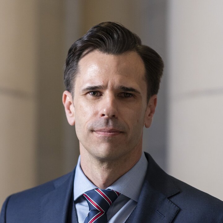 Man in formal suit with striped tie, in a softly lit indoor setting.